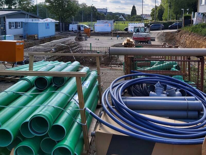 Eine Baustelle mit gestapelten grünen Rohren, blauen Kabeln und Fahrzeugen im Hintergrund.