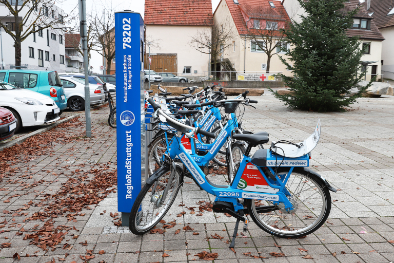 Es wird das Regiorad Station gezeigt. Dies steht an einer blauen Säule. Das Fahrrad selbst ist auch blau. Es steht auf einem gepflasterten Boden. Links sind einige Autos zu sehen.