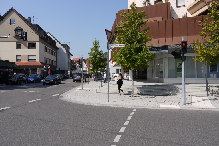 Es ist eine Ampel zu sehen, welche rot anzeigt. Im Hintergrund sind Häuser und Autos. Eine Person läuft Richtung Straße.