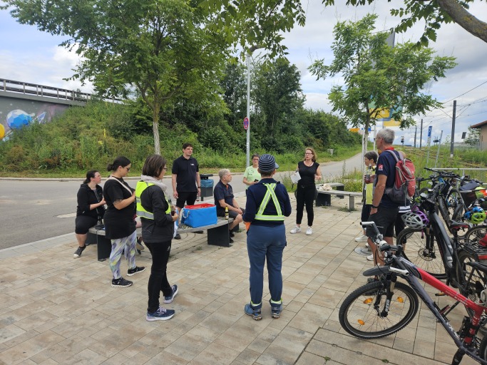 Foto auf dem mehrere Menschen zu sehen sind. Rechts stehen Fahrräder. Die Menschen sitzen und stehen.